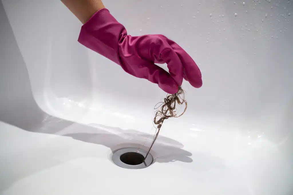 Hand of woman in pink rubber glove taking off hair clump from water drain, blocked pipe in bath., Bathroom Drain.